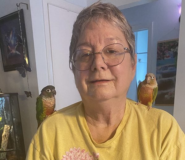 Short haired woman closeup of face with a cinnamon green cheek conure on both shoulders