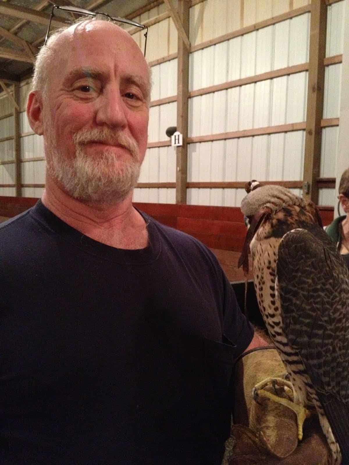 Mitch Rezman with short beard and hooded peregrine falcon on left arm